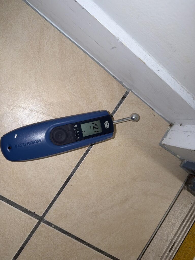 Ball-probe moisture meter checking a tiled floor beside the wall during a water damage response